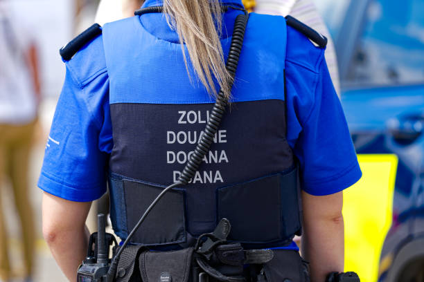 Back view of a customs officer in uniform wearing a vest with radio equipment, indicating border control duty.