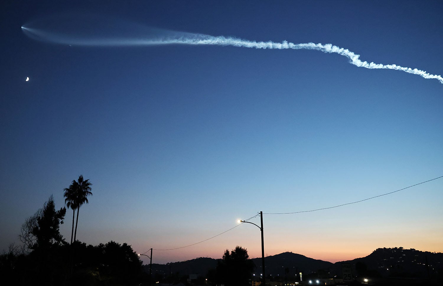 A rocket streaks across the sky at dusk, appearing as just a tiny pinprick of white light with a long, snaking white chemtrail extending behind it, nearly horizontal across the blue-to-pink gradient of the sky. Palm trees and telephone wires are visible at the bottom of the image.