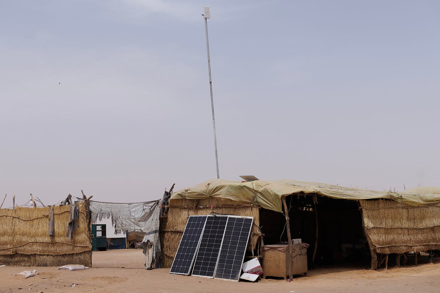 A hut sits on a sandy lot, with solar panels propped up beside it and a tall pole with a Starlink device on top of it. Other small huts are visible beyond it.