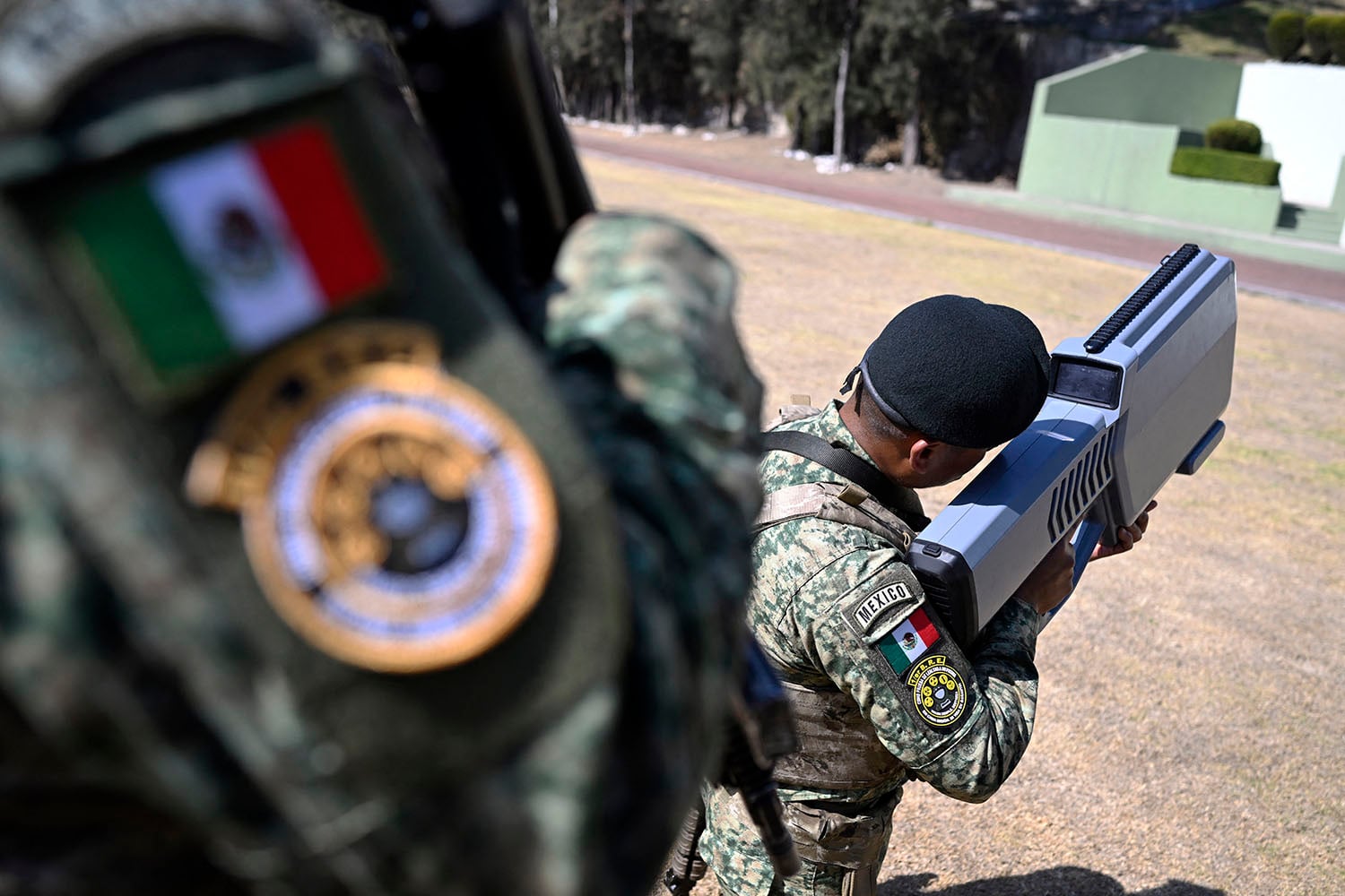 The arm and patches of a soldier are visible in the foreground, partly obscuring another another soldier in a camouflage uniform beyond, facing away as they hold a rectangular missile-launcher-like device.