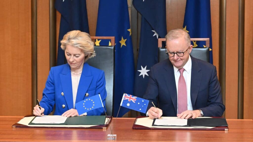 Ursula von der Leyen and Anthony Albanese sitting next to each other at a desk, signing documents. There are Australian and EU flags behind them.