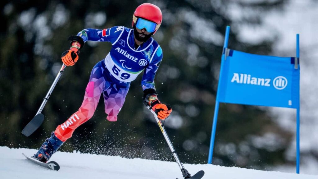 A para-alpine skier with prosthetic legs races down a snowy slope during a slalom event, wearing a red helmet, reflective goggles, and a blue racing suit.