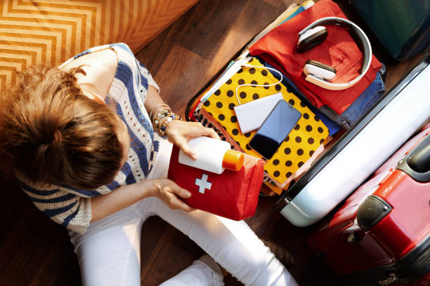 Woman packing a first aid kit into an open suitcase with travel items like headphones and a phone.