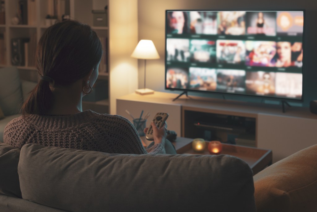 A woman relaxing on a couch, using a remote control to choose a show from the TV menu.