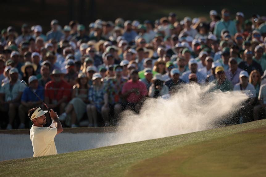 A golfer hits a ball out of a bunker, with a wave of sand captured flowing through the air from the shot.