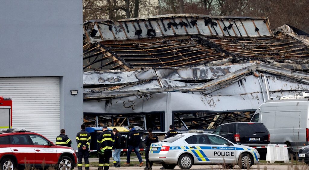 Police officers and a firefighters stand in front of the burned production hall in Pardubice, Czech Republic, March 20, 2026.