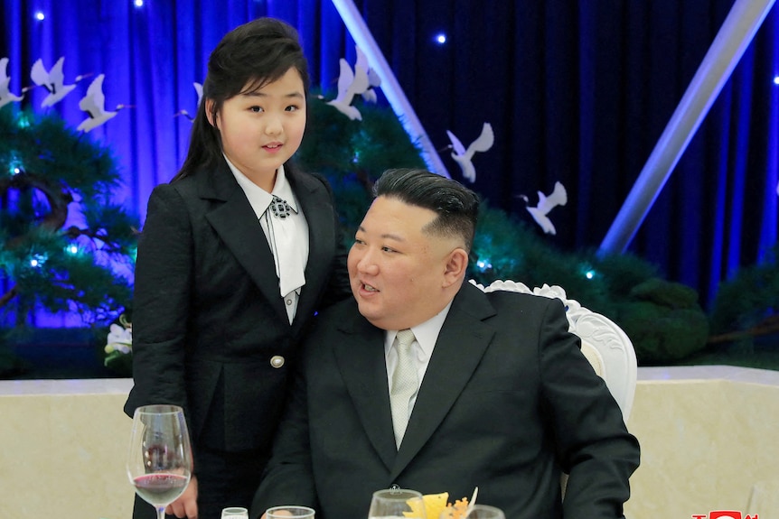 A girl stands next to a man sitting down.