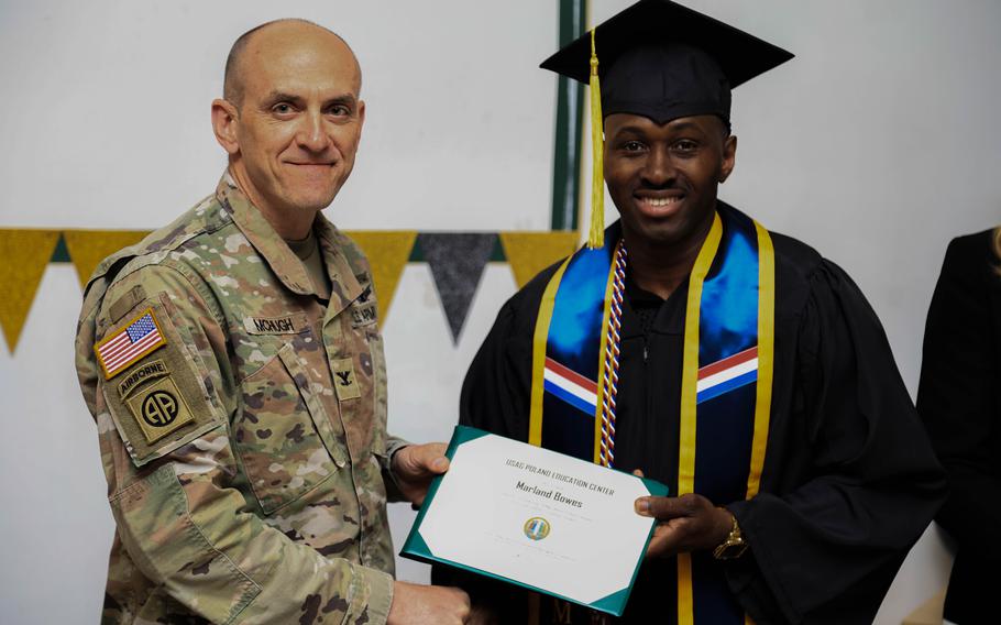 A U.S. Army colonel presents a graduation certificate to a soldier in cap and gown at USAG Poland’s education ceremony.