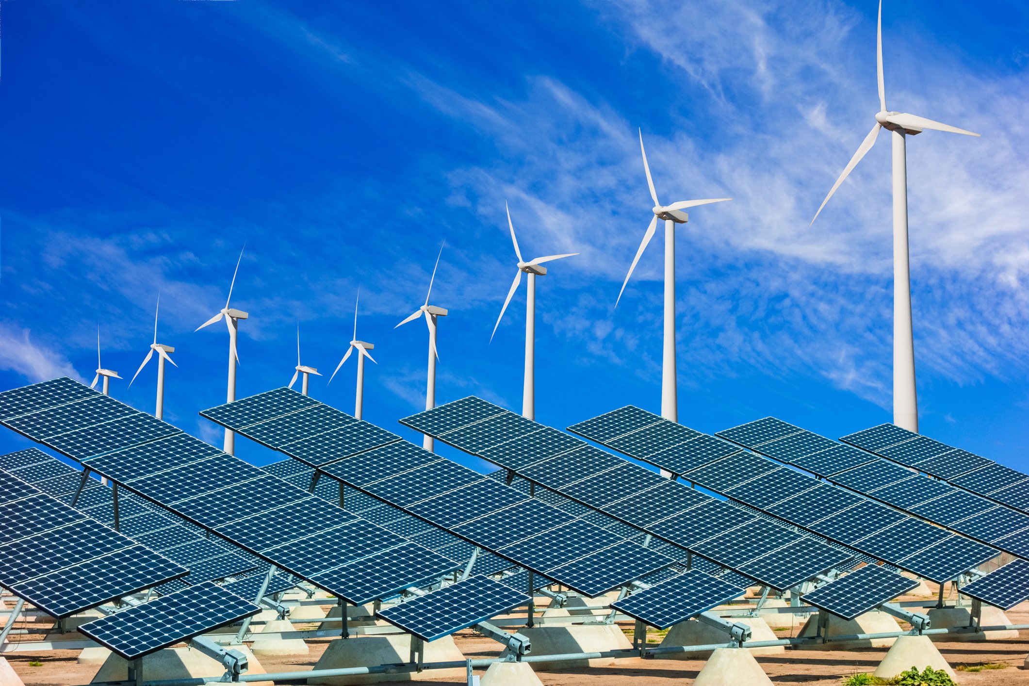 In front of a bright blue sky, multiple wind turbines and solar panels are visible.