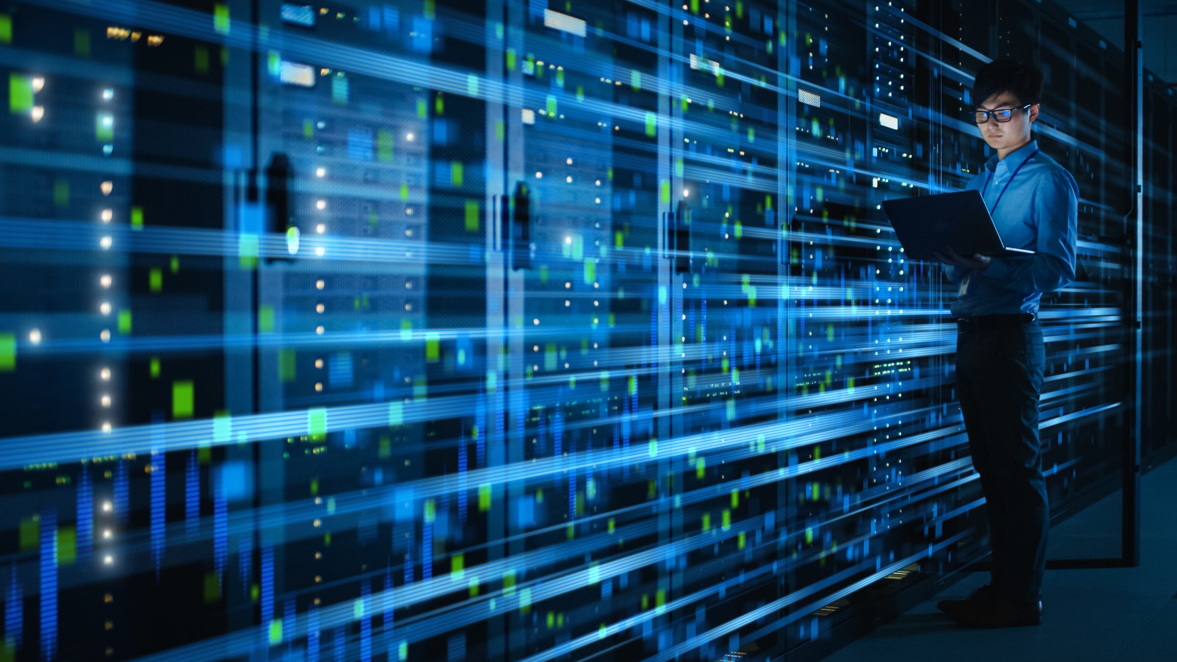 A person holding a laptop in front of racks of servers.