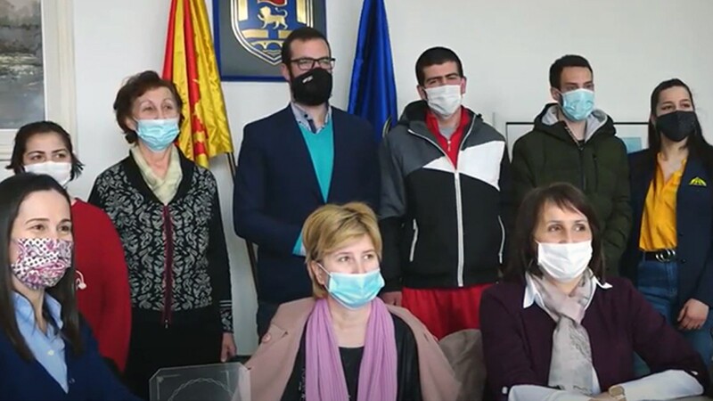 Six people standing behind three women seated, all wearing masks. On the wall in the background, a seal between two flags. 