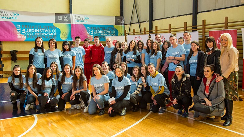 A group photo of athletes, coaches and supporters on two rows in a gym standing in front of Special Olympics banners.