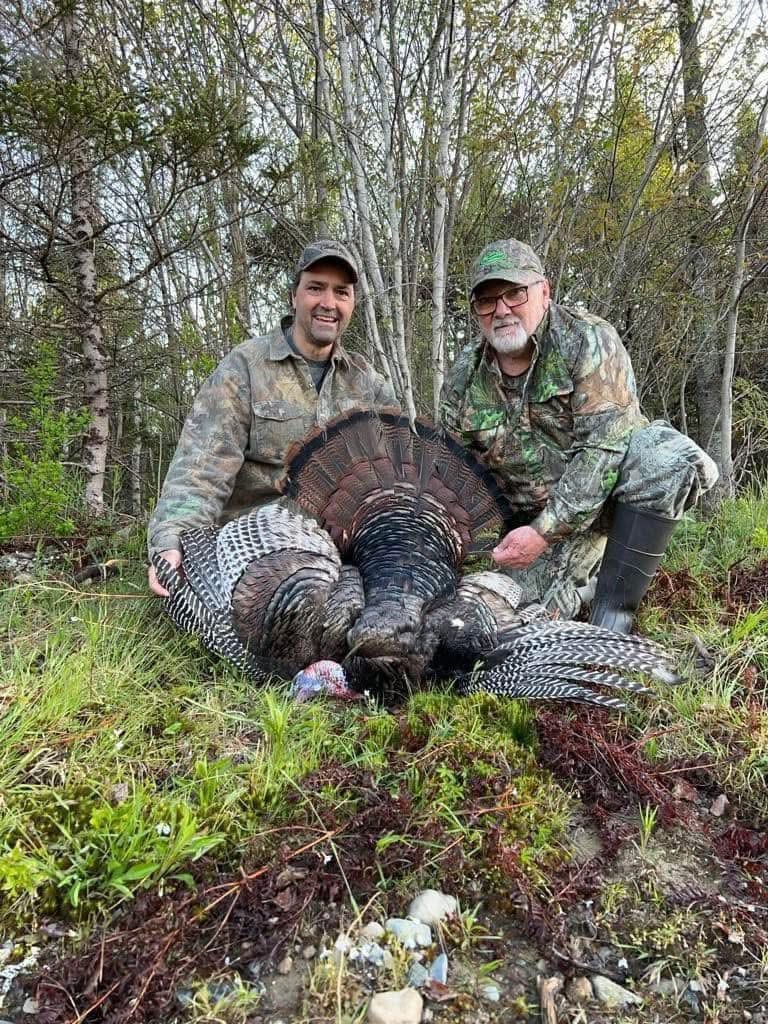 Two men in camo holding a dead turkey