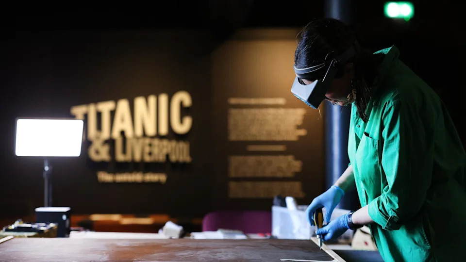 A female worker works on preserving a painting rolled out on a table in front of her. She wears goggles, a green overall and blue latex gloves. The sign Titanic and Liverpool can be seen behind.