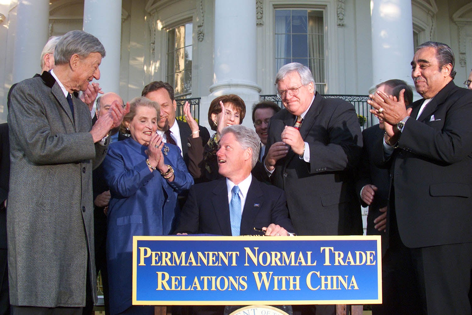 A photograph of Bill Clinton sitting at a desk outdoors, smiling and looking toward a group of people. In front of him is a blue sign with gold lettering that reads, "Permanent Normal Trade Relations With China." Several men and women in formal attire stand behind him, clapping.