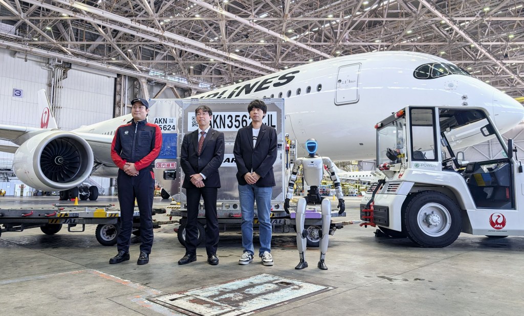 A humanoid robot and three Japan Airlines staff stand in front of a JAL plane in a hangar.