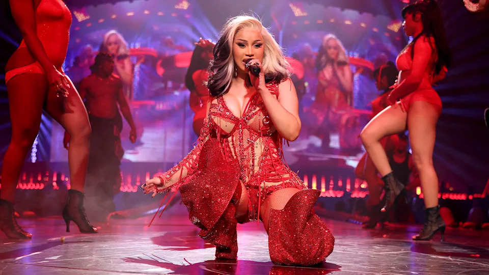 Cardi B in a red outfit sings on stage with dancers in the background. The stage is lit with vibrant lights and displays.