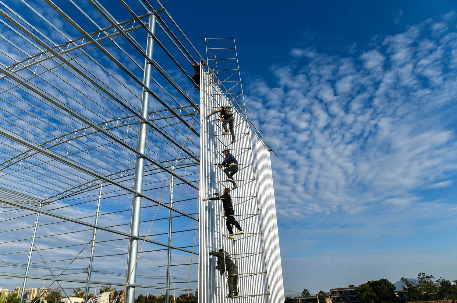 A wide shot showing workers on a scaffolding structure, attaching large white corrugated panels to a tall, silver metal frame of a building under construction. The background features a bright blue sky with wispy white clouds and a glimpse of a city skyline in the distance.