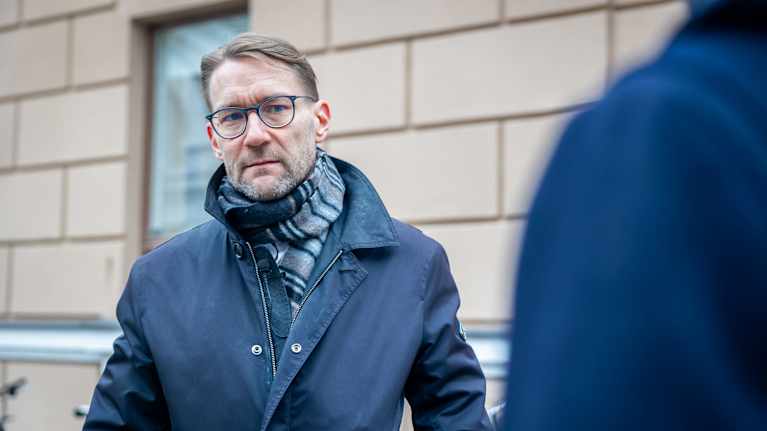 A man with a stubbly beard, glasses and a dark scarf and coat stands outside a beige building with a serious expression.