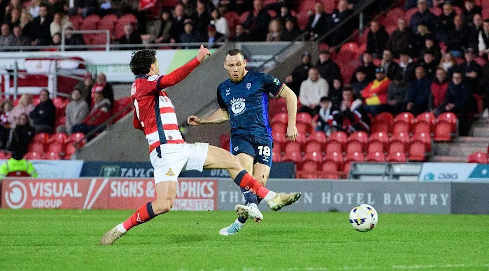 Ben House, wearing a dark blue football kit, takes a shot towards goal as a player wearing red and white stripes slides in for a tackle. Red seats, supporters and advertising boards are in the background.