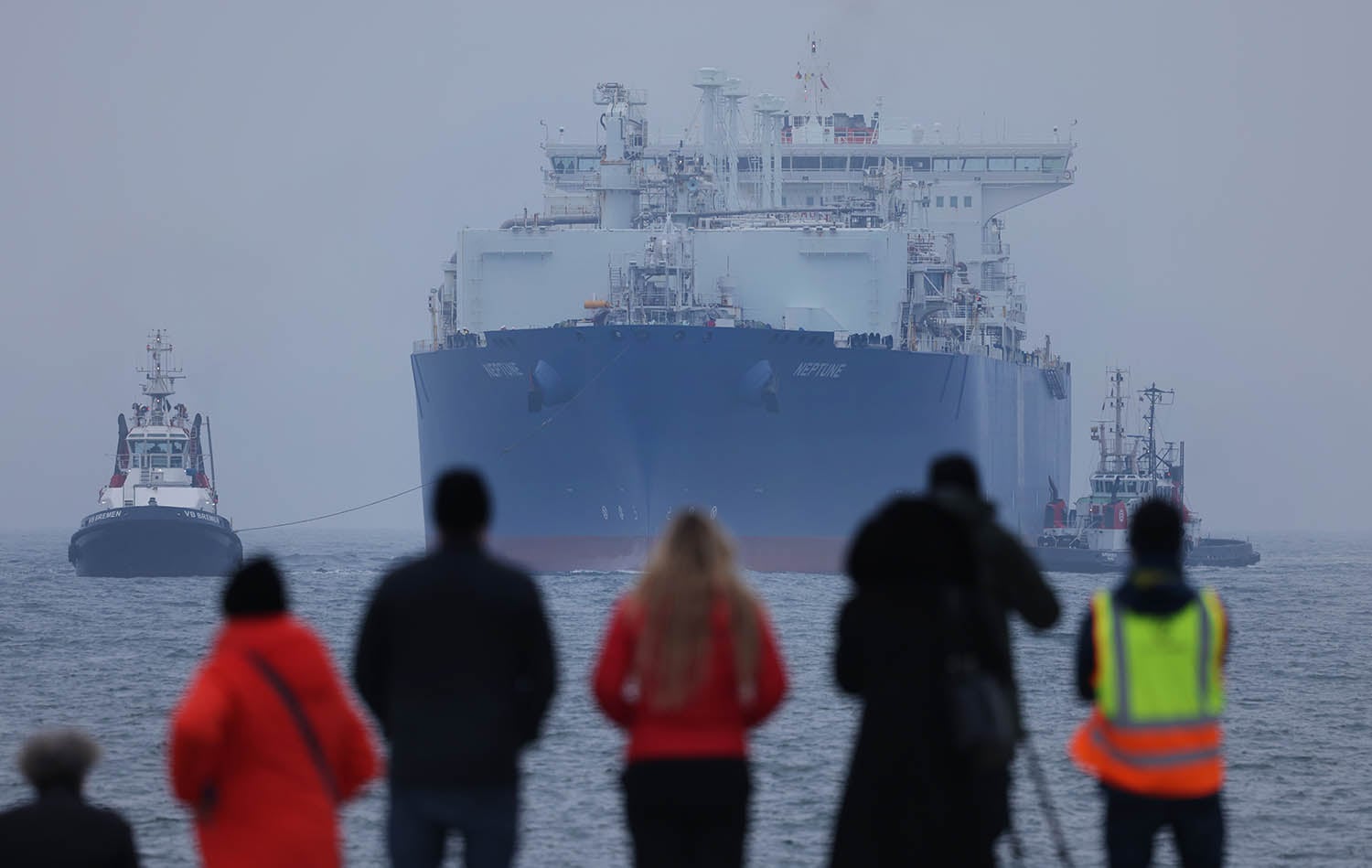 A large blue and white tanker ship named "NEPTUNE" is being guided through calm, misty waters by two smaller tugboats. In the foreground, several people are seen from behind, watching the ship from a shoreline.