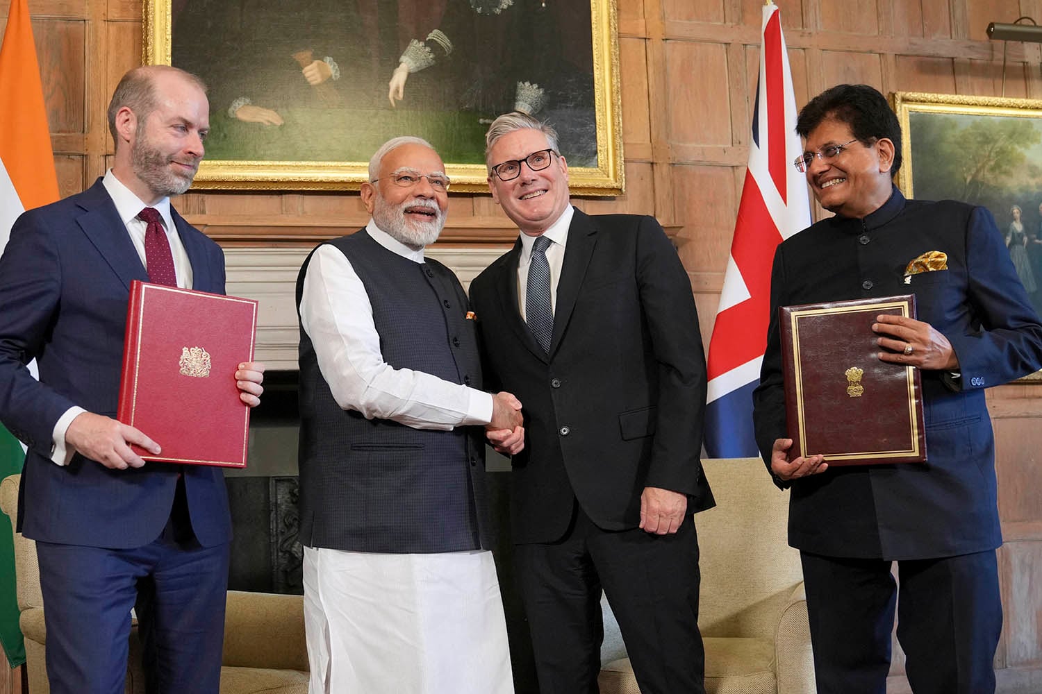 A group of four men stand together in a wood-paneled room. In the center, two men are shaking hands, while the men on either side hold large, formal leather-bound folders. The flags of India and the United Kingdom are visible in the background.