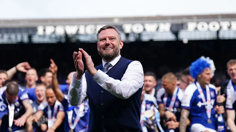 Mark Ashton is wearing a white shirt, a navy blue knitted waistcoat and a striped tie. He is smiling and applauding, with Ipswich Town footballers celebrating behind him.