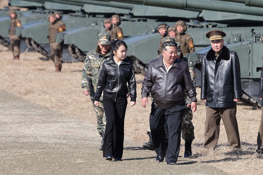 A girl and several men wear leather jackets as they walk alongside tanks.