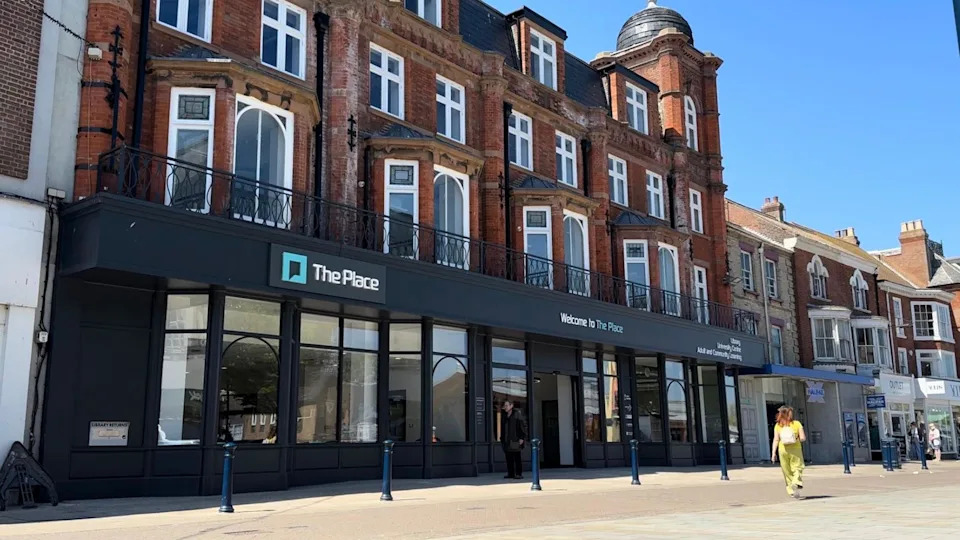 A large red-bricked building with a dark grey frontage on its lower floor is in view as a woman wearing a yellow outfit walks past in Great Yarmouth's Market Place