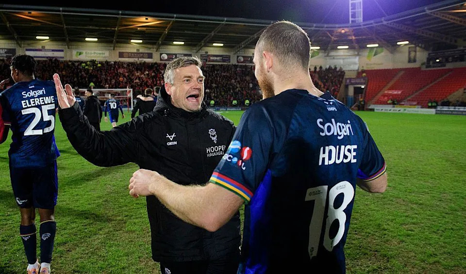 Two people stand on a football pitch after a match, facing each other with arms raised as they talk. One man is wearing a black padded jacket while the other wears a dark blue playing kit with the number 18 on the back. Additional players, red stadium seating and a crowd are in the background.