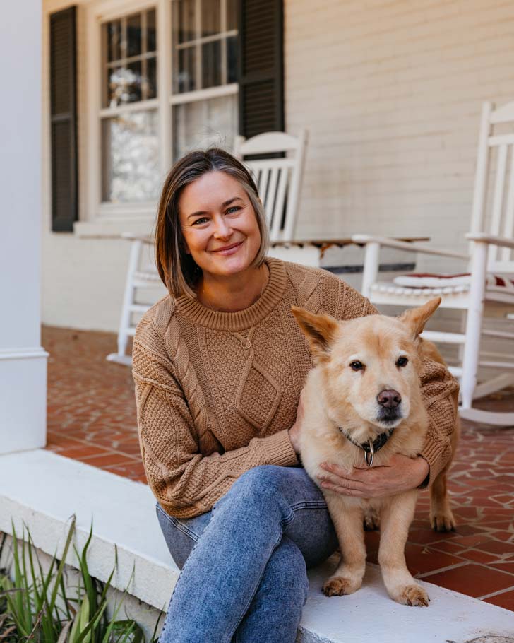 Katharine K. Wilkinson sits on her front porch, in front of two white rocking chairs. Next to her is a tan dog with a graying muzzle