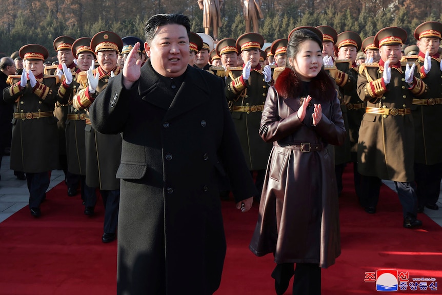 Kim Jong Un walks in front of soldiers in uniform cheering him, with his daughter in a brown coat alongside him