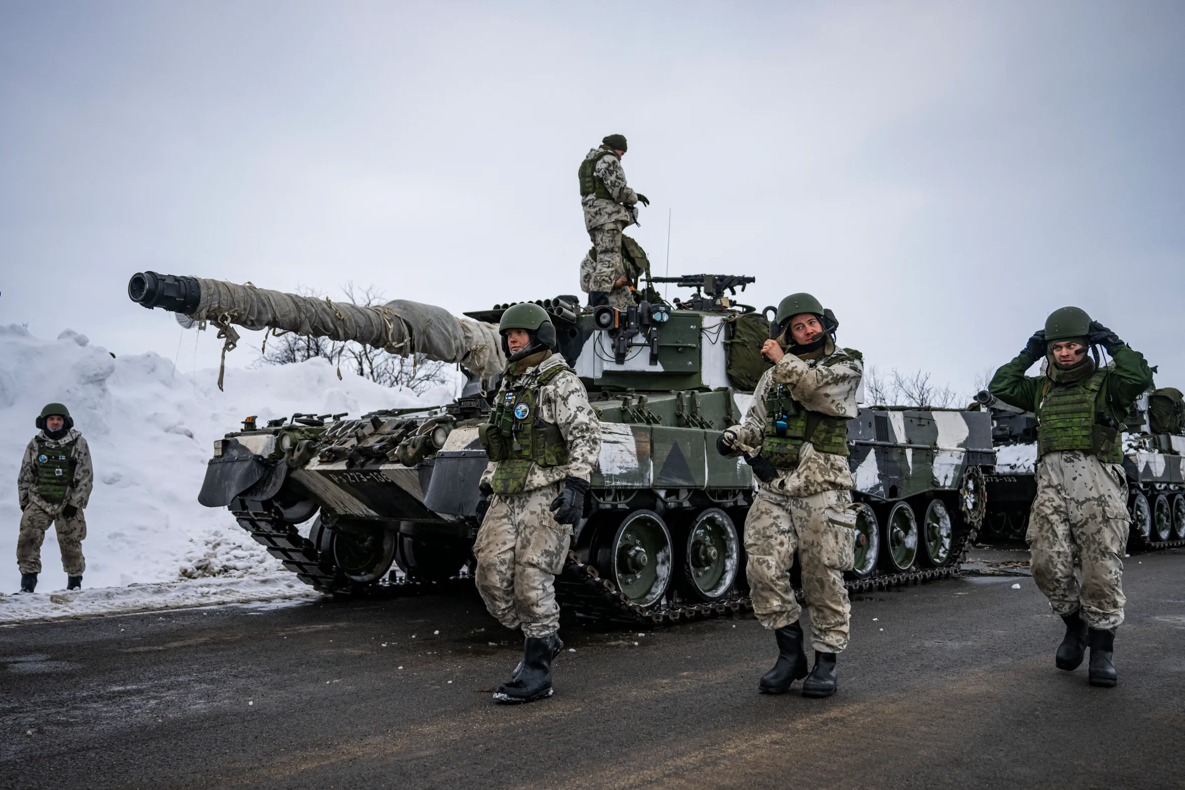 Finnish soldiers in camouflage uniforms and helmets stand on a road next to a Leopard 2A6 tank with a wrapped barrel in a snowy, outdoor setting.