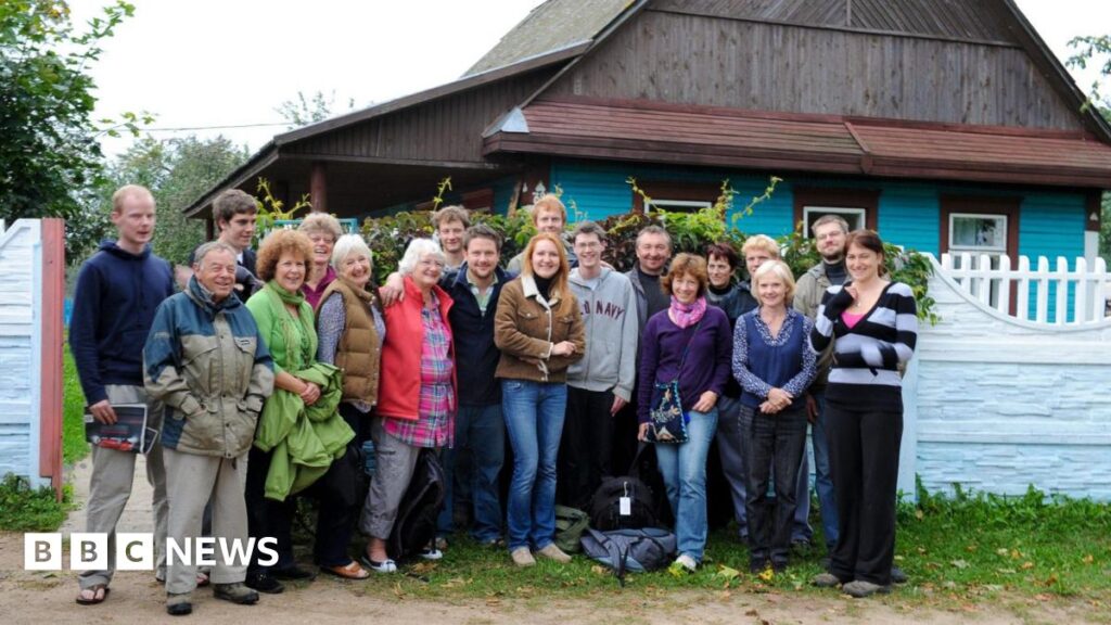 A group of people wearing jackets, jumpers and scarves stands outdoors in front of a wooden building and a pale blue fence in a rural setting, photographed in 2010.