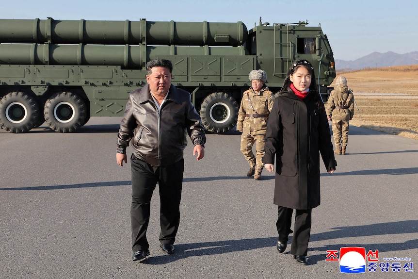 A man an a young woman walk side-by-side with a large truck with cylinders in the background