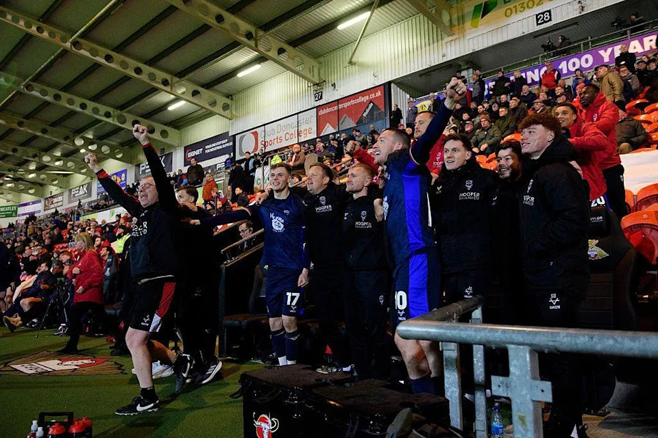 Football players and coaching staff stand along the touchline in a stadium, raising their arms in celebration. The group wears dark blue kits and black coats, with seated spectators and advertising boards behind them.