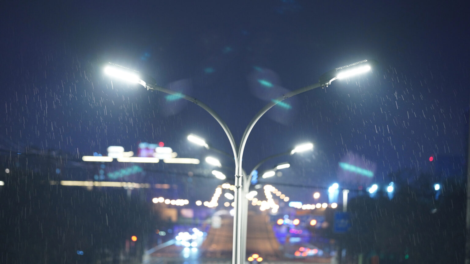 Streetlights illuminate a rainy night, with blurred city lights in the background creating a moody atmosphere.