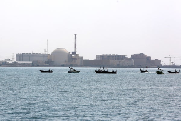 The low, gray buildings of a power plant seen with Persian Gulf waters and boats in the foreground