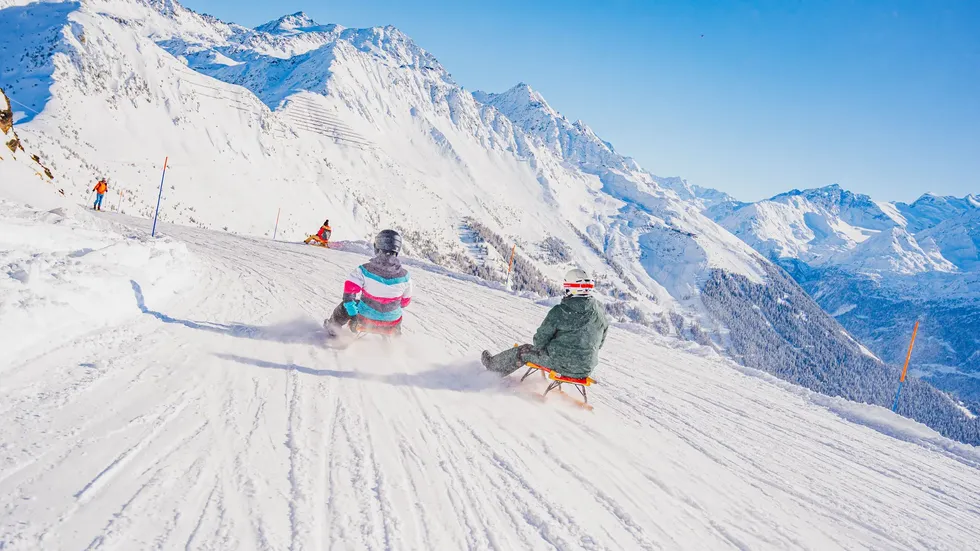 two individuals sledding down a snowy slope with mountains in the background