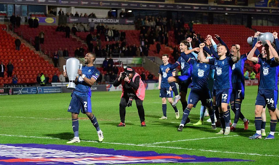 Football players in dark blue kits walk across the pitch carrying a large, silver inflatable trophy. Several players are raising their arms as they walk past a pitchside photographer. Red stadium seating, spectators and advertising boards are in the background under floodlights.