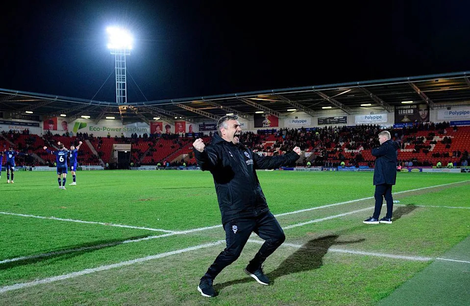 Michael Skubala, the head coach of Lincoln City, celebrates at the final whistle. He is wearing a black coat and tracksuit bottoms. Several players are in the background with their arms raised in celebration. They are standing on a green football pitch surrounded by football stadium seating. An illuminated floodlight is set against a dark sky.