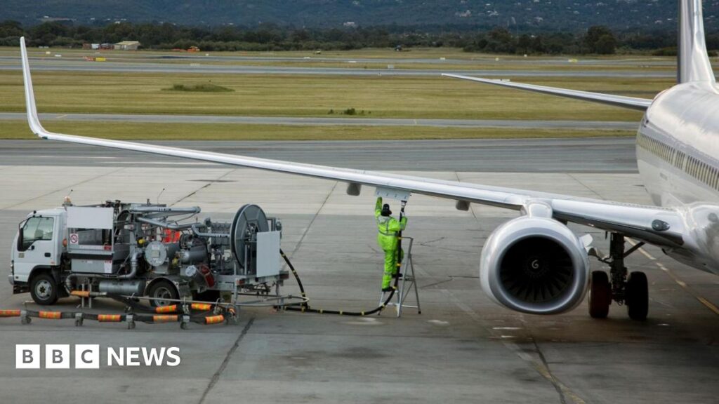 A man in a fluro yellow suit refuels an airplane sitting on an airport tarmac.