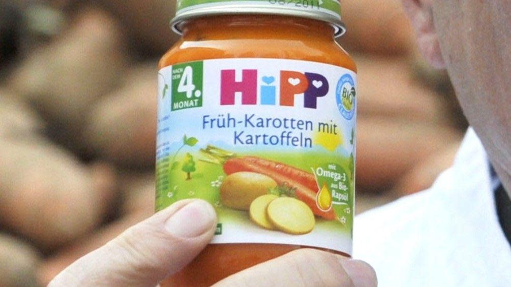 A man holds a jar of HiPP carrot and potato baby food, with potatoes stacked in the background.