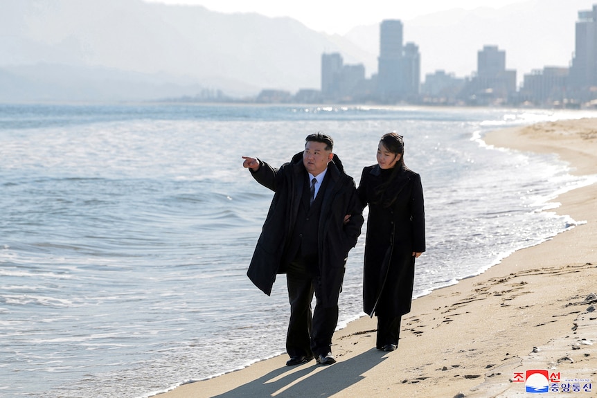  Kim Jong Un and Kim Ju Ae walk along a beach with a city in the backgrounds. 