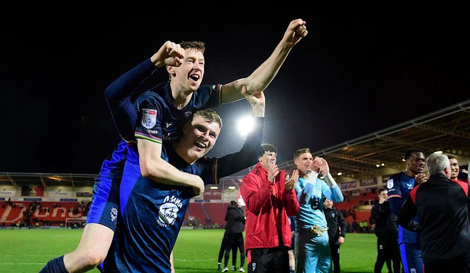 Two football players in dark blue kits celebrate on the pitch, with one player lifted on another’s back and raising an arm. Other players and staff stand nearby, clapping.