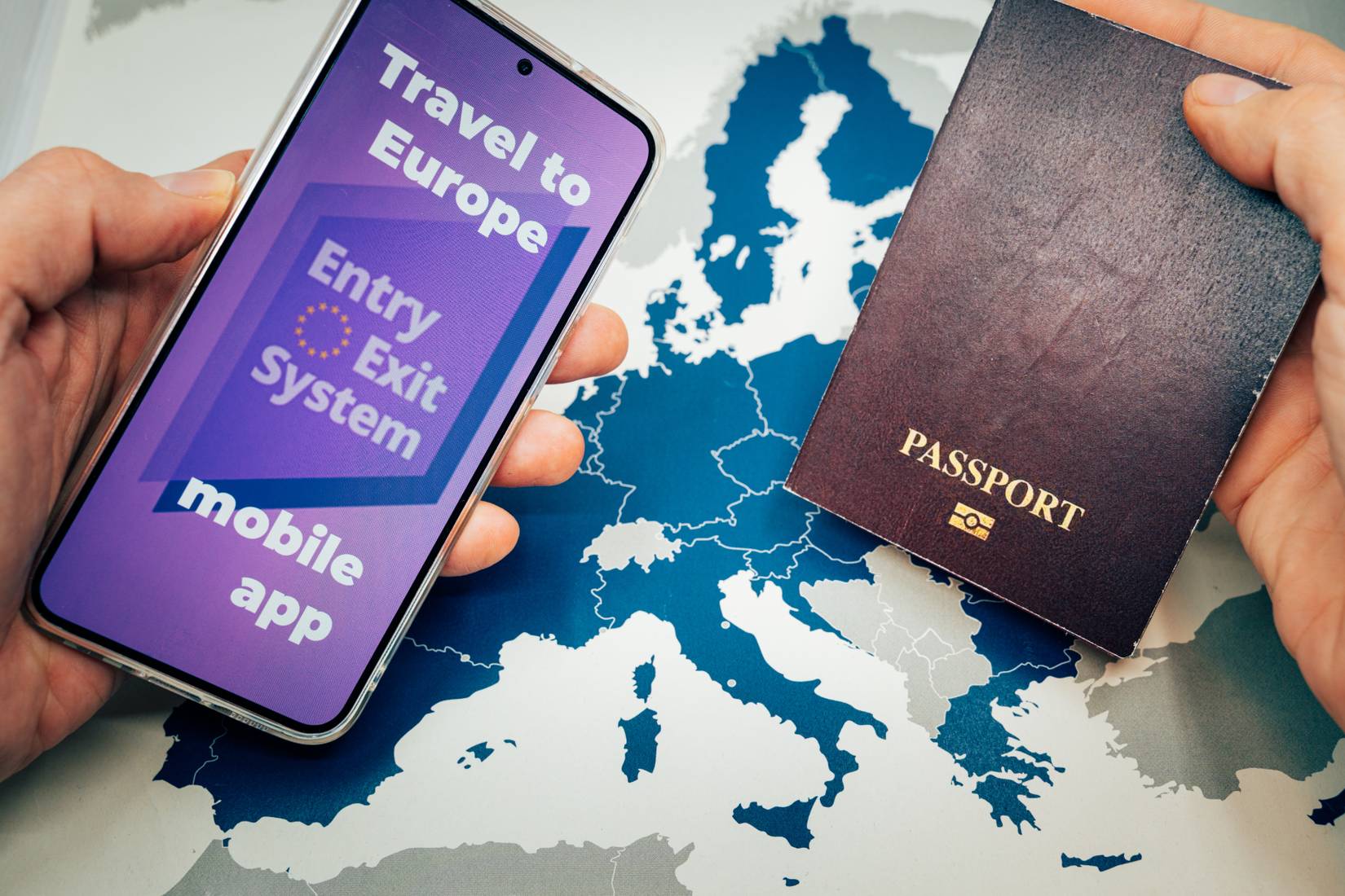 Hands placing a passport and a smartphone with the Entry Exit System (EES) mobile app on a map of Europe, a new requirement for non-EU travelers entering the Schengen Area starting in 2025
