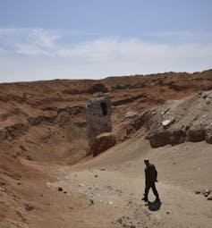 silhouetted man walks in foreground of dusty landscape with a large metallic object poking out of crater