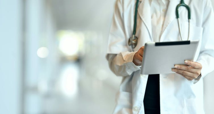 A doctor in a white coat with a stethoscope around their neck holds an electronic tablet.