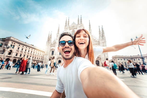 Couple smiling and taking a selfie in a busy plaza with a large cathedral behind them.
