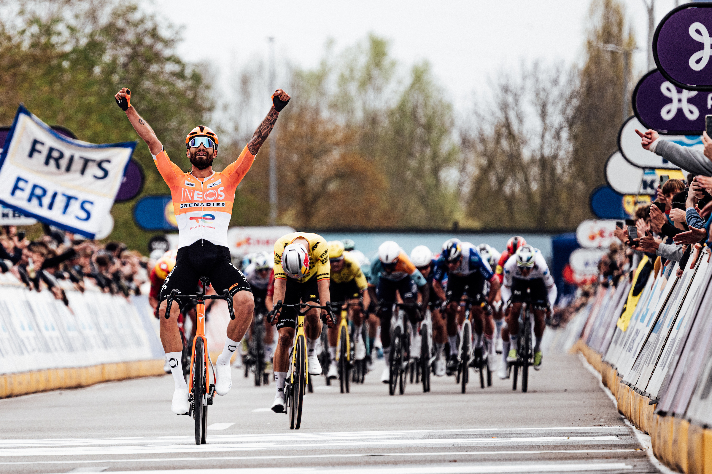 A photo of Filippo Ganna celebrating winning Dwars door Vlaanderen in front of the sprinting peloton, with Wout van Aert dropping his head in disappointment. To the right of the shot, you can see an arm reaching over the barrier, giving he middle finger